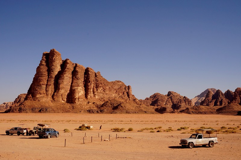 A rock formation known as "The Seven Pillars of Wisdom" in Wadi Rum, Jordan is massive as it dwarfs the pickup trucks in the foreground