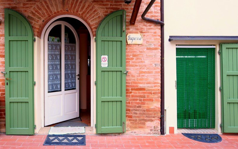 Charming entrance of an Italian agriturismo in Emilia-Romagna, featuring rustic red brick walls, green wooden shutters, and a welcoming open door, capturing the authentic countryside hospitality travelers experience when staying in rural Italy