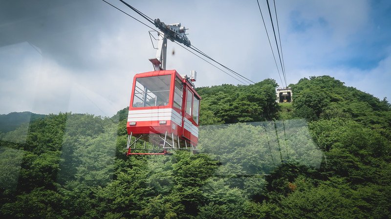 Bright red Akechidaira Ropeway cable car gliding above lush green forested mountains in Nikko, Japan, with panoramic views unfolding under a partly cloudy sky