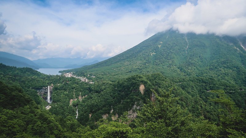 Panoramic view from the Akechidaira Ropeway Lookout showing Kegon Falls plunging into a forested gorge, Lake Chuzenji glimmering in the distance, and lush green mountains surrounding Nikko, Japan, under a partly cloudy blue sky
