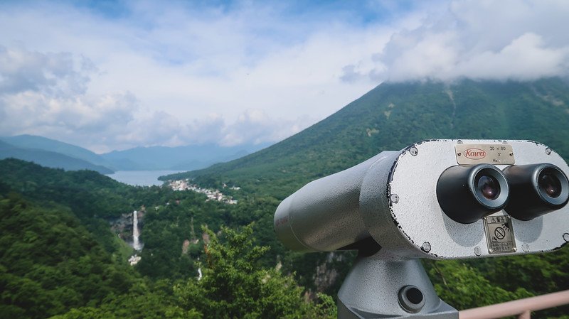 Coin-operated binoculars at the Akechidaira Ropeway Observation Point overlooking Kegon Falls cascading down the cliffs and Lake Chuzenji nestled among lush green mountains in Nikko, Japan, under a partly cloudy sky