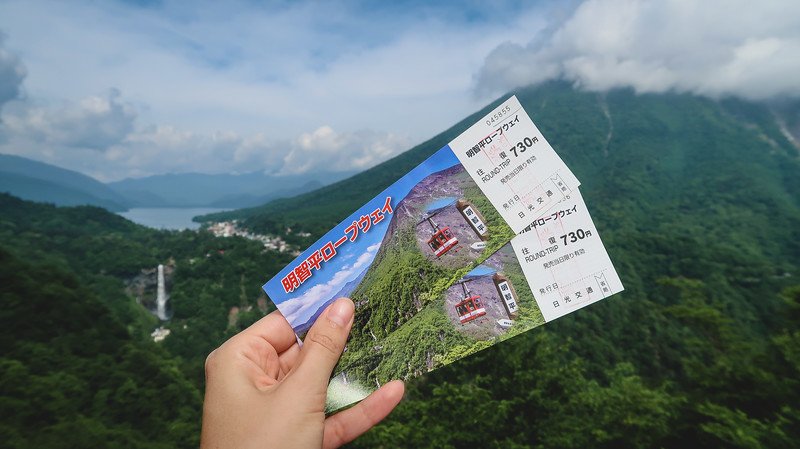 Hand holding two colorful Akechidaira Ropeway tickets with mountain and cable car illustrations, overlooking Lake Chuzenji and Kegon Falls surrounded by lush green mountains in Nikko, Japan, under a partly cloudy sky
