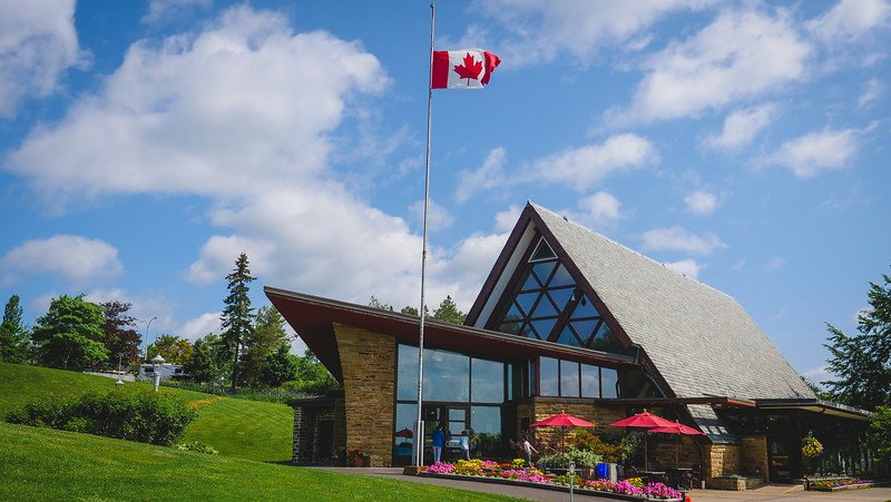 A tall Canadian flag waves in the breeze outside the striking A-frame building of the Alexander Graham Bell National Historic Site in Baddeck, Nova Scotia, surrounded by vibrant flower beds, green lawns, and blue summer skies during a road trip along the Cabot Trail in Cape Breton
