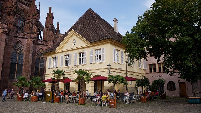 Visitors relaxing on the sunny outdoor patio of Alte Wache wine bar, a charming historic building located beside Freiburg Cathedral in Freiburg im Breisgau, Germany