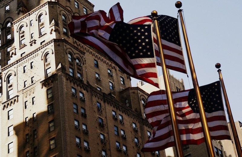 Multiple American flags wave proudly in the breeze against the backdrop of a historic high-rise building in Midtown Manhattan, New York City, capturing a classic urban scene with strong patriotic symbolism and architectural detail