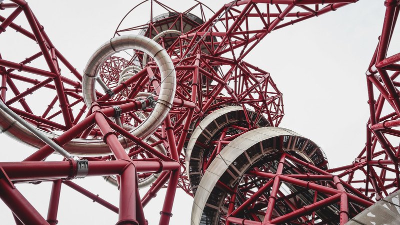 Looking up at the twisting red steel structure of the ArcelorMittal Orbit in London’s Queen Elizabeth Olympic Park, showcasing the dramatic loops of the world’s longest tunnel slide wrapped around the iconic landmark’s framework