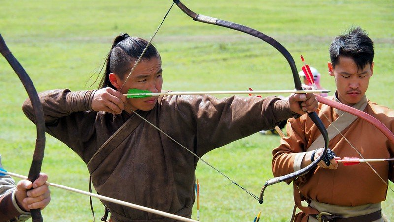 Archery games at the World Nomad Games with a Kyrgyz man with long hair and traditional attire about to shoot the arrow from the bow