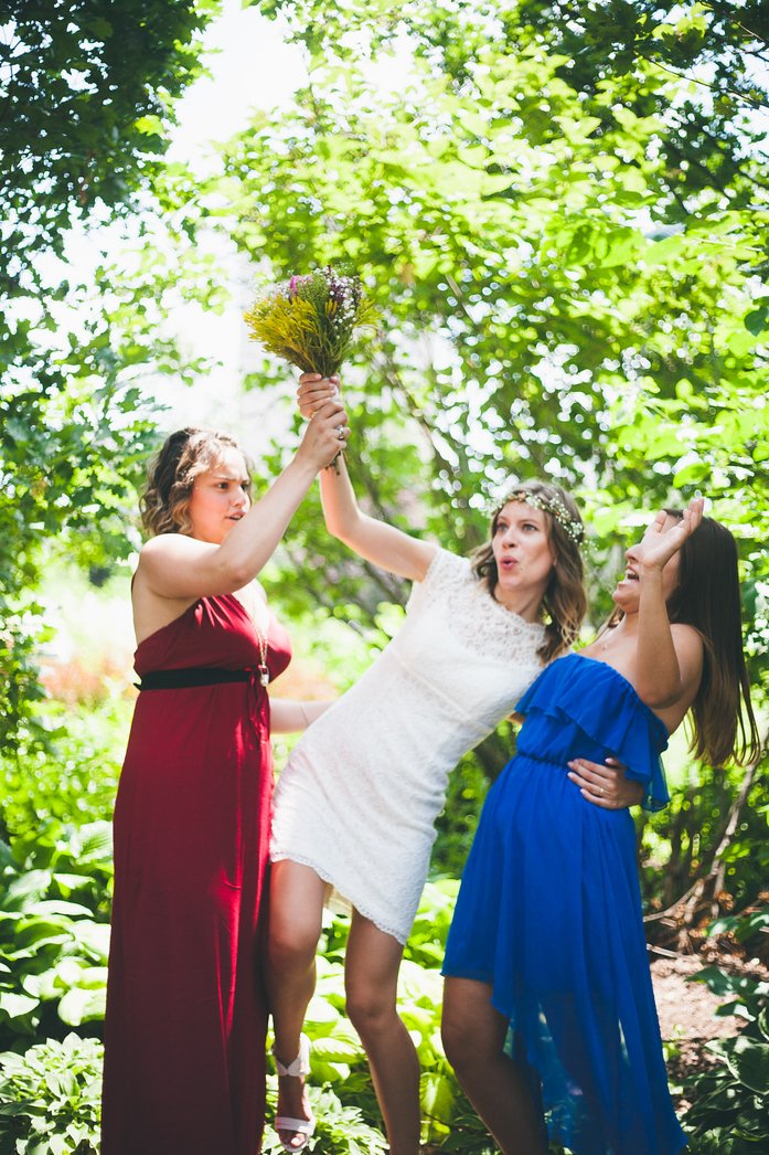 Candid wedding moment with Audrey Bergner in a lace dress and flower crown, joined by bridesmaids in red and blue dresses, laughing as their heels sink into the garden path while Ashley saves the bouquet during Samuel and Audrey’s summer wedding celebration