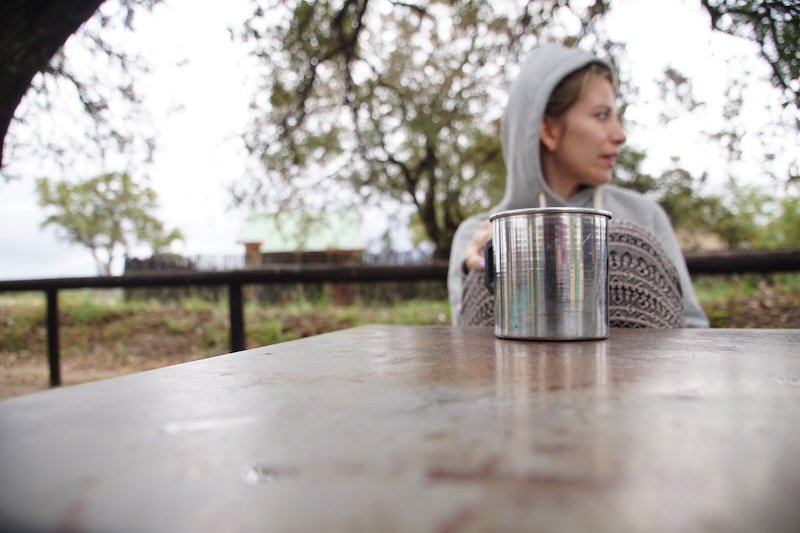 That Backpacker Audrey Bergner wearing a cozy hoodie to stay warm while sipping morning coffee from a tin mug at a rustic campsite during a chilly Kruger National Park safari in South Africa