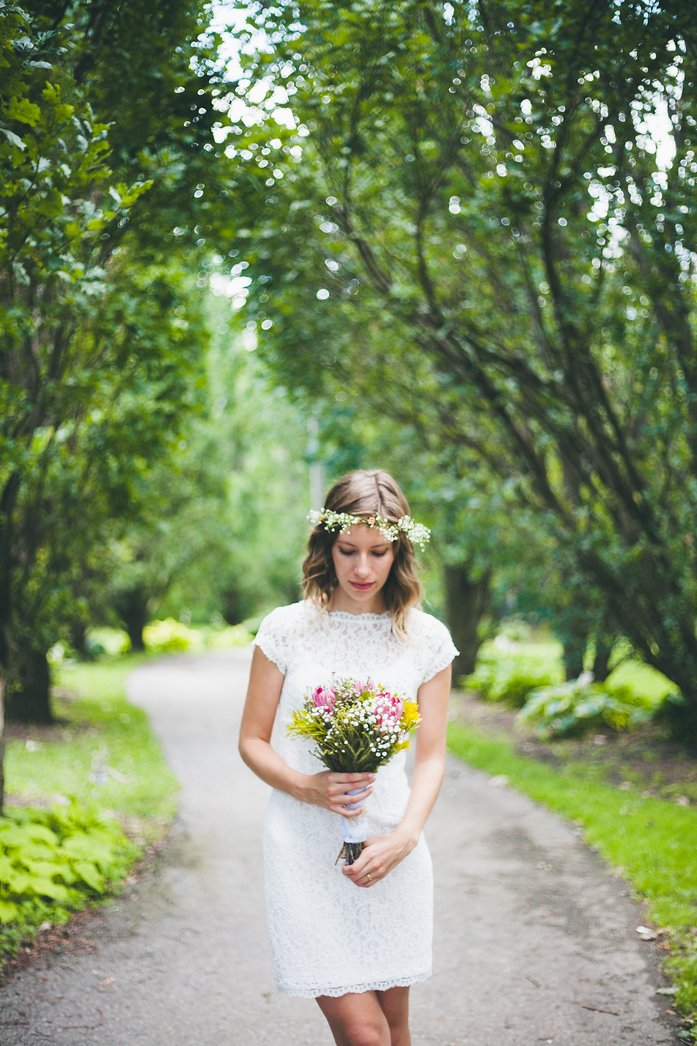 Audrey Bergner of That Backpacker stands gracefully on a tree-lined path in Brampton, Ontario, wearing a delicate lace wedding dress and floral crown of baby’s breath, holding a vibrant bouquet of South African proteas during her summer wedding