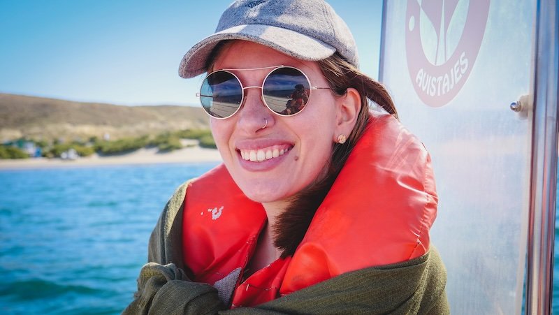 Audrey Bergner of That Backpacker smiles brightly while wearing sunglasses and a life jacket during a scenic boat tour in Puerto Pirámides, Península Valdés, Argentina. The calm blue waters and coastal landscape frame the sunny adventure perfectly