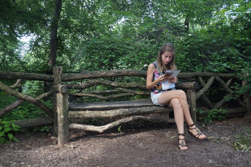 Audrey Bergner of That Backpacker sits on a rustic wooden bench surrounded by lush greenery in New York City’s Central Park, carefully studying a map during a summer trip. The peaceful woodland setting captures a quiet travel moment in the city