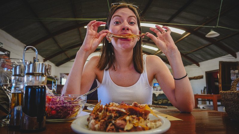 Audrey Bergner of That Backpacker enjoying a traditional Patagonian lamb (cordero) lunch at Estancia San Lorenzo on Península Valdés, Argentina. Seated in a rustic dining hall, she playfully holds up a lamb bone, capturing the authentic countryside food experience
