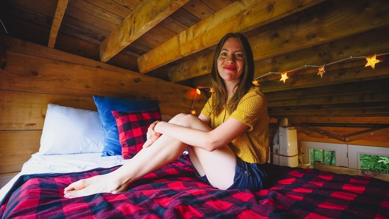 Audrey Bergner of That Backpacker sitting cross-legged on the bed in the cosy loft of a tiny cabin Airbnb in Glen Huron, Ontario, Canada. The warm wood interior, string lights, and red-and-black plaid blanket create a rustic, inviting atmosphere
