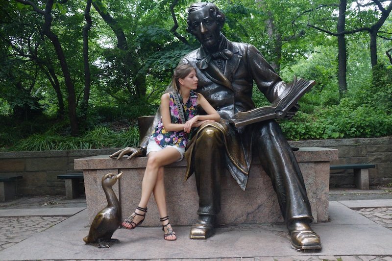 Audrey Bergner of That Backpacker poses beside the Hans Christian Andersen statue in Central Park, New York City, during a summer trip. Sitting on the bench next to the famous author and storyteller, she playfully interacts with the iconic sculpture