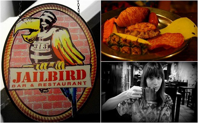 Audrey Bergner of That Backpacker enjoying breakfast at Jailbird Restaurant in Hotel Katajanokka, Helsinki, Finland, with a plate of assorted pastries, cheese, fruit, and meats, alongside the colorful parrot-themed restaurant sign in the historic jail hotel