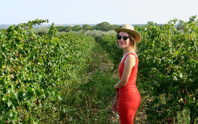 Audrey Bergner of That Backpacker enjoying a sunny afternoon at La Vinyeta vineyard in Costa Brava, Spain, wearing a red dress and straw hat while walking through lush grapevines, capturing the charm of Spanish wine country during a winery tour.