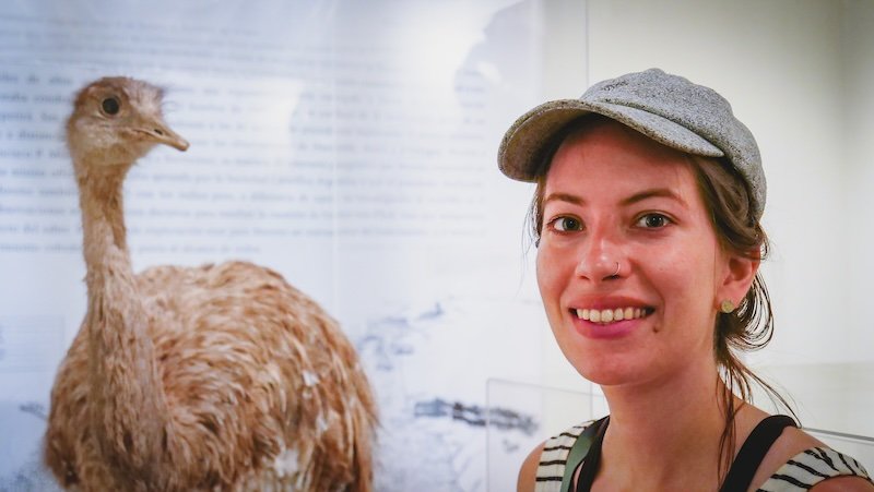 Audrey Bergner of That Backpacker smiles beside a taxidermy rhea on display inside a museum in Puerto Madryn, Patagonia, Argentina, highlighting the region’s wildlife exhibits and cultural attractions that make this coastal city a fascinating stop