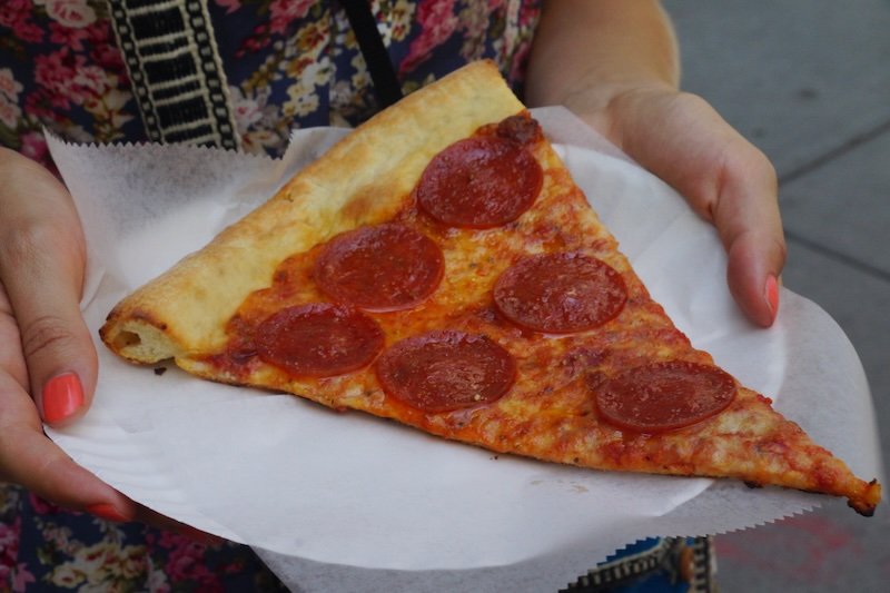 Audrey Bergner of That Backpacker holds a classic New York pepperoni pizza slice on a paper plate during her summer trip to NYC. The thin crust and generous cheese are typical of iconic New York-style pizza, making it a must-try food experience