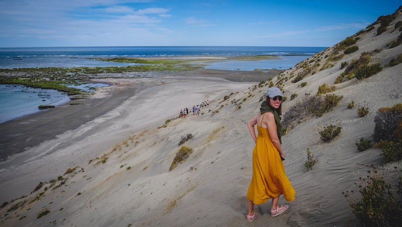 Audrey Bergner of That Backpacker hikes down a sandy dune overlooking the dramatic coastline of Península Valdés in Patagonia, Argentina. The view stretches across tidal flats and the Atlantic Ocean, showcasing the region’s unique coastal landscapes and natural beauty
