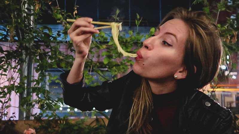 Audrey Bergner of That Backpacker slurping noodles with chopsticks at Koi Ramen in Pop Brixton, London, surrounded by lush greenery and evening lights, enjoying the vibrant street food culture during a 24 hours in London food adventure