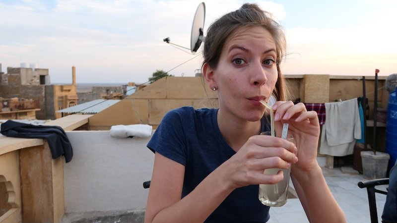 Audrey Bergner of That Backpacker sipping a refreshing drink on a rooftop terrace at sunset in Jaisalmer, Rajasthan, India, with sandstone buildings and desert views in the background