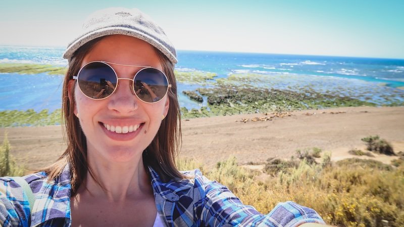 Audrey Bergner of That Backpacker takes a cheerful selfie wearing a hat and sunglasses while visiting Punta Norte on Península Valdés, Argentina. Behind her, the rugged Patagonian coastline, turquoise waters, and sea lion colony create a stunning backdrop