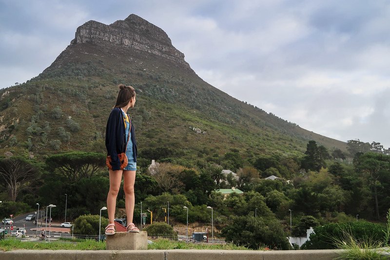Audrey Bergner from That Backpacker standing on a low ledge gazing up at Signal Hill in Cape Town, surrounded by lush greenery and urban streets at the mountain’s base