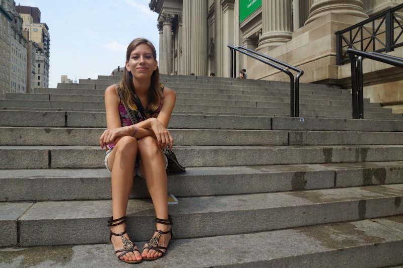 Audrey Bergner of That Backpacker sits relaxed on the iconic steps of the Metropolitan Museum of Art in New York City, with the museum’s grand neoclassical columns rising behind her on a warm summer day, capturing a classic NYC travel moment