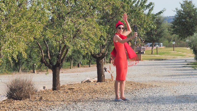 Audrey Bergner of That Backpacker vlogging selfie-style at La Vinyeta winery in Costa Brava, Spain, wearing a vibrant red dress and traditional Catalan scarf, capturing her passion for storytelling during a grape stomping and wine tasting experience