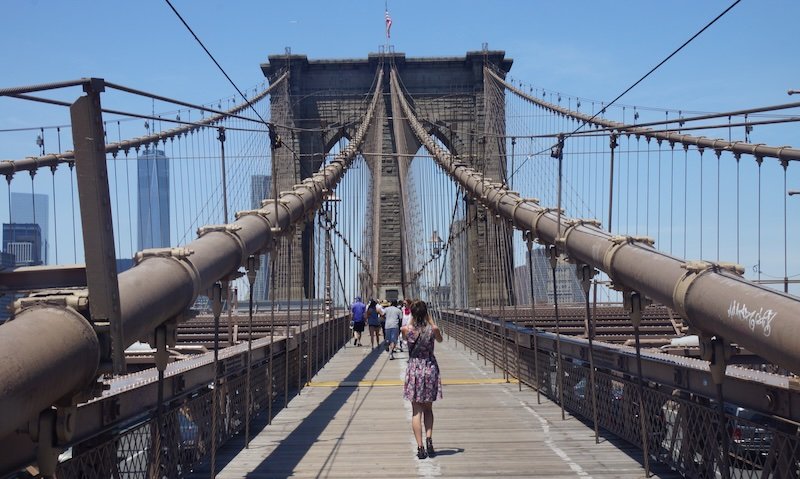 Audrey Bergner of That Backpacker walks along the wooden pedestrian path of the Brooklyn Bridge in New York City on a sunny summer day, capturing iconic travel photos with the Manhattan skyline in the background. A classic NYC sightseeing experience