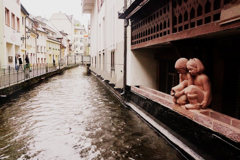 Charming canal known as a Bächle flowing between historic buildings in Freiburg im Breisgau, Germany, featuring two small child statues perched along the water’s edge