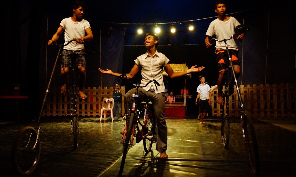 Acrobatic performers riding tall bicycles and doing theatrical stunts under bright stage lights during a vibrant show at Phare Ponleu Selpak Circus in Battambang, Cambodia, showcasing contemporary Cambodian circus arts and storytelling.