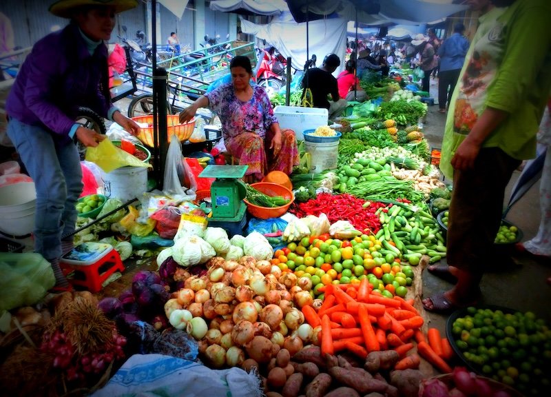 A vibrant rural market scene in Battambang, Cambodia, with local vendors selling colorful fresh produce like carrots, limes, onions, chilies, and herbs during our one-month Cambodia travel itinerary