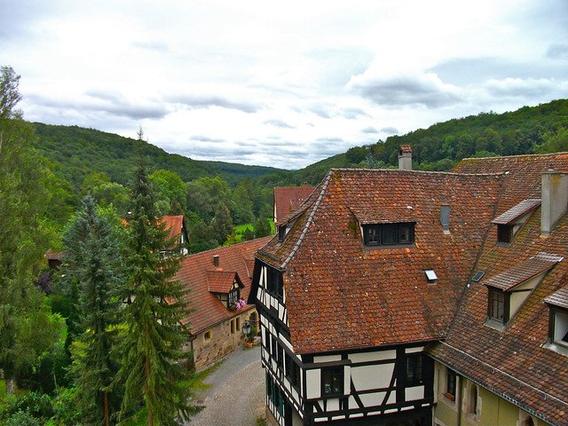 Scenic view of Bebenhausen Monastery and village in Germany, featuring half-timbered houses, red tiled rooftops, cobblestone lanes, and lush forested hills in the background under a cloudy sky, creating a timeless medieval setting