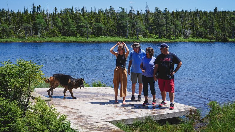 Nomadic Samuel and Audrey Bergner That Backpacker's family stands on a small wooden dock by the calm blue waters of Benjie Lake in Cape Breton Highlands National Park, surrounded by evergreen forest, while their dog Togo explores the edge of the dock on a sunny summer day along the Cabot Trail in Nova Scotia