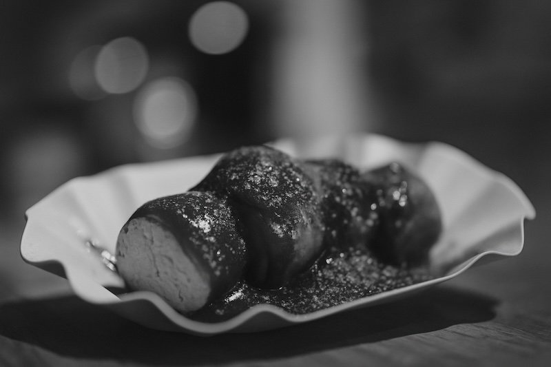 A black and white macro photograph of Berlin currywurst served in a scalloped paper tray. The sliced sausage is covered in curry ketchup and sprinkled with curry powder, captured from a creative off-center angle with strong bokeh in the background for depth