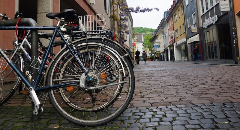 Row of parked bicycles on a cobblestone street in Freiburg im Breisgau, Germany, with colourful buildings and distant hills visible in the background