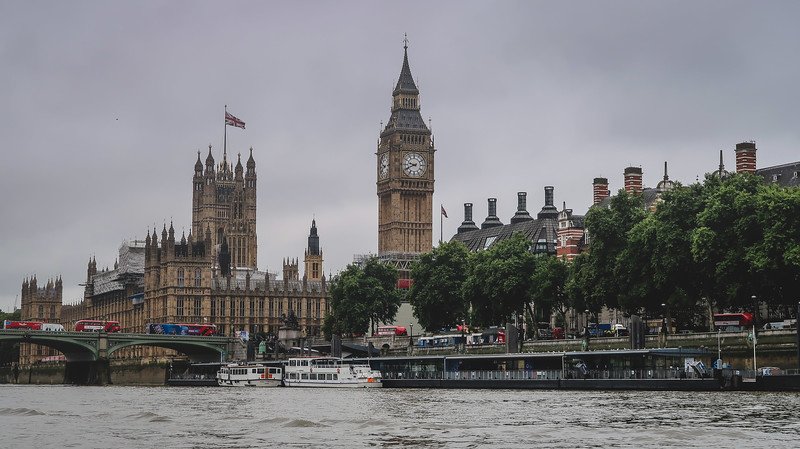 The Palace of Westminster and Big Ben rise above the River Thames in London, with red double-decker buses crossing Westminster Bridge and boats docked along the riverside, showcasing one of the city’s most iconic and quintessential views
