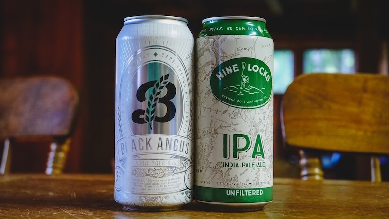 Two cans of local craft beer — Black Angus IPA from Big Spruce Brewing in Cape Breton and Nine Locks IPA from Nine Locks Brewing in Dartmouth, Nova Scotia — sit on a wooden table, highlighting local flavours to try while road tripping the Cabot Trail