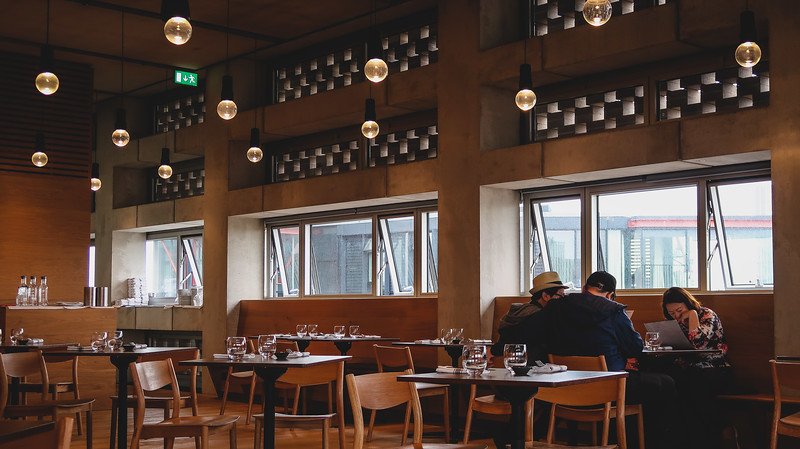 Diners sit inside the restaurant at the Blavatnik Building in Tate Modern, London, surrounded by modern wooden furnishings, soft hanging lights, and large windows that create a stylish yet relaxed setting for meals with a city view
