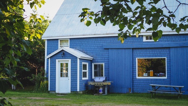 A charming blue-painted barn with white trim, a picnic table, and a barbecue grill sits nestled among leafy trees, offering a cozy and unique barn stay experience along the scenic Cabot Trail in Cape Breton, Nova Scotia