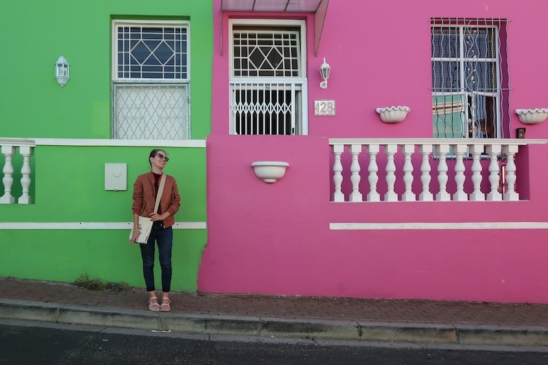 That Backpacker Audrey Bergner standing in front of the vibrant green and hot pink houses of the Bo-Kaap neighbourhood in Cape Town, South Africa, known for its colourful facades and rich cultural heritage