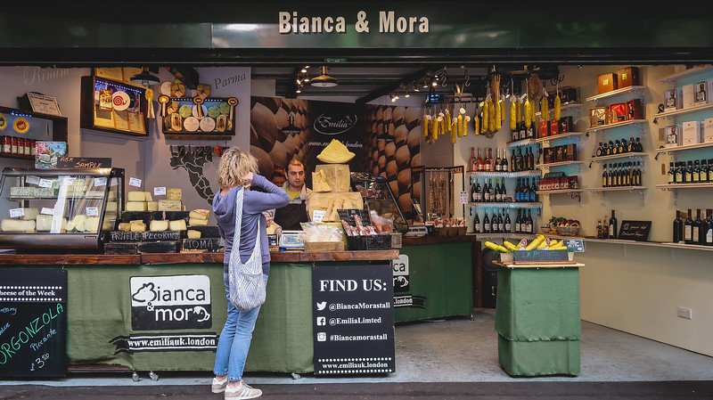 A customer shops at Bianca & Mora in Borough Market, London, where wheels of cheese, cured meats, and bottles of wine fill the stall, showcasing Italian specialties and artisanal flavors that make this market a paradise for food lovers