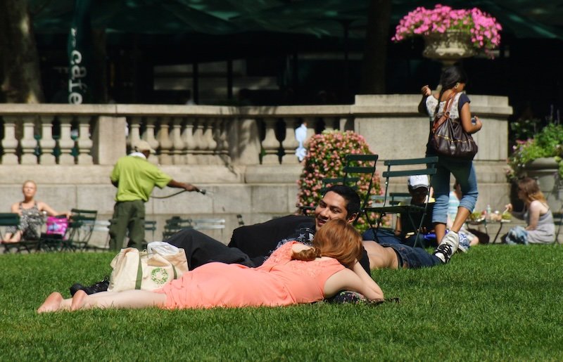 People lounging and relaxing on the lawn at Bryant Park in Midtown Manhattan, New York City, on a warm summer day. A couple reclines on the grass surrounded by flowers and park chairs, capturing the local vibe of enjoying green spaces in the heart of NYC