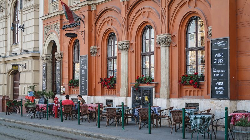 Outdoor café in Budapest with red and orange architecture, arched windows, and tables lined with checkered cloths and flowers in bloom, inviting visitors to enjoy Hungarian cuisine, wine tastings, and the lively atmosphere of the city