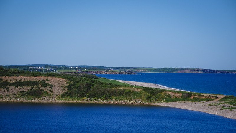 A panoramic view of rugged coastline and narrow sandspit jutting into the Atlantic Ocean, framed by vivid blue waters and rolling green hills along the scenic Cabot Trail in Cape Breton, Nova Scotia