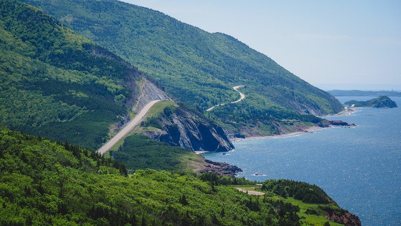Scenic aerial view of the winding Cabot Trail hugging the rugged coastline of Cape Breton Island in Nova Scotia, with lush green forested hills meeting the deep blue waters of the Atlantic Ocean