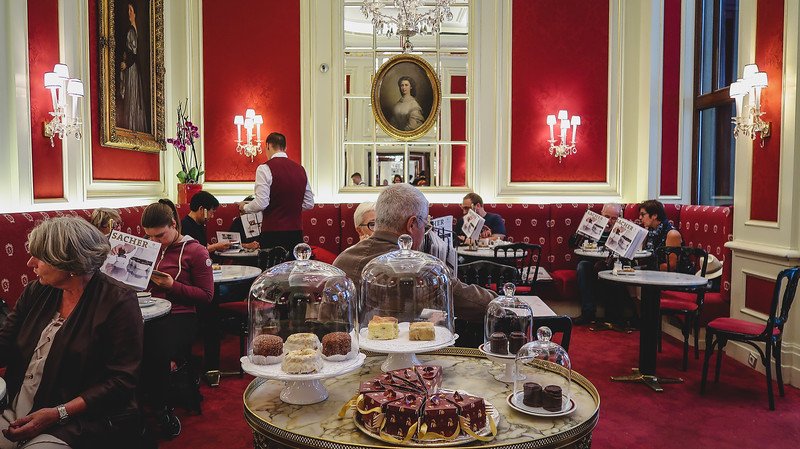 Elegant red and gold interior of Café Sacher Wien in Vienna, Austria, with patrons enjoying coffee and the famous Sachertorte surrounded by chandeliers and classical portraits Elegant red and gold interior of Café Sacher Wien in Vienna, Austria, with patrons enjoying coffee and the famous Sachertorte surrounded by chandeliers and classical portraits