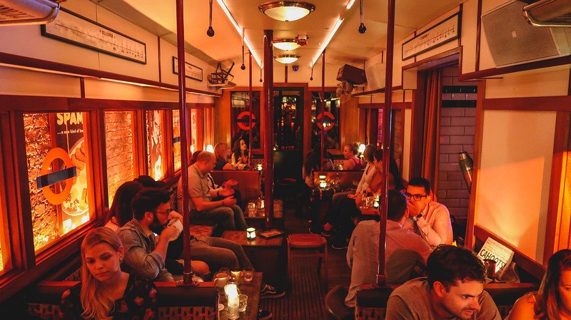 Guests seated inside Cahoots, a 1940s-themed underground bar in London designed to look like a vintage tube carriage, with retro posters, warm candlelit tables, and nostalgic décor creating a lively speakeasy atmosphere reminiscent of wartime London